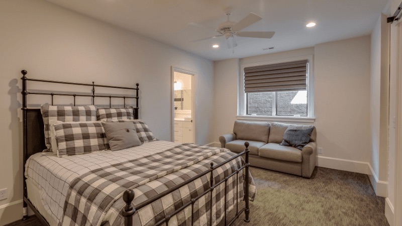 Basement guest bedroom with plaid bedding, a loveseat, and a bright egress window